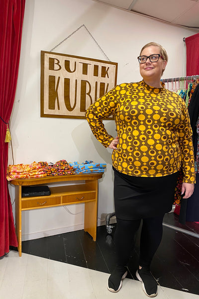A person in glasses and a yellow Kubik sweater pop by BUTIK KUBIK stands smiling near a wooden snack table. A Butik KUBIK sign is visible behind them, alongside clothing racks.