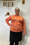 Wearing glasses and the Kubik sweater popsicle by BUTIK KUBIK in bright orange and pink, a person poses confidently with one hand on their hip before a wall displaying "KUBIK," pairing the vibrant knit top with a black skirt.