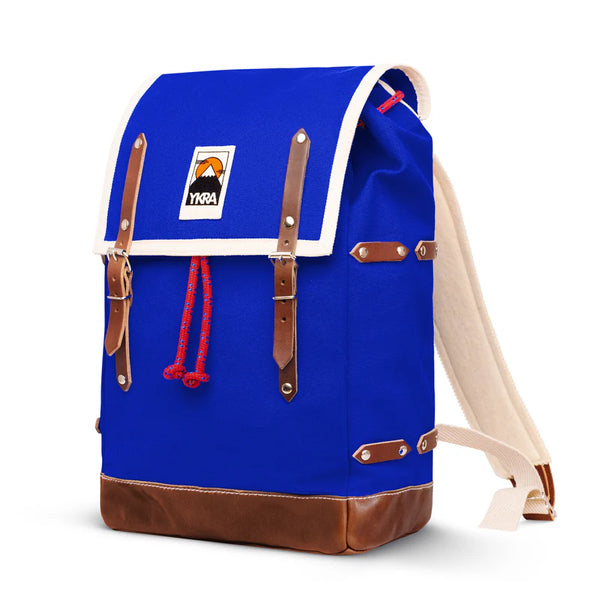 The BUTIK KUBIK YKRA Matra is a blue canvas backpack with brown leather straps and base, white trim on the flap, red rope drawstring, and a front patch featuring a mountain design, evoking classic vintage mountaineering style.