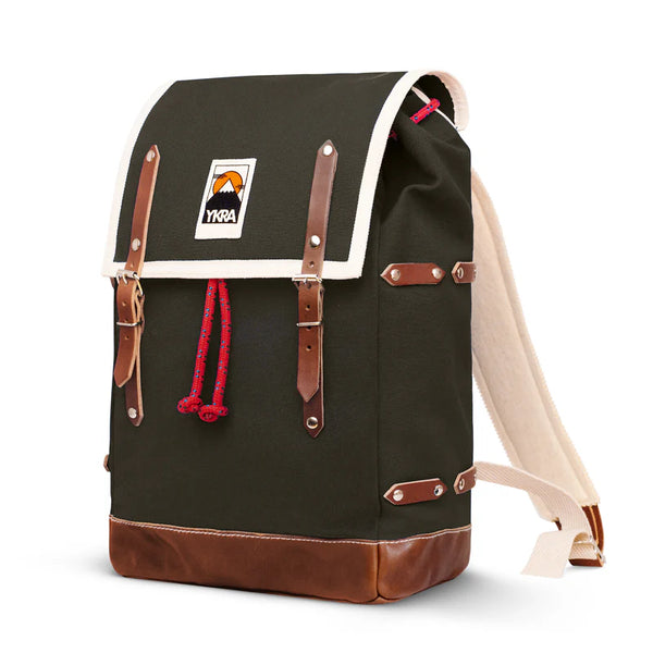 The BUTIK KUBIK YKRA Matra (green) is a vintage-inspired backpack crafted from dark green cotton canvas with brown leather straps, cream trim, a red drawstring, a mountain logo patch on the flap, and a reinforced brown leather base.