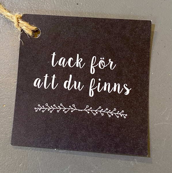 The Tack för att du finns card from BUTIK KUBIK features a black square tag with a jutesnöre loop, white handwritten Swedish text meaning thank you for existing, and a branch design. It has an elegant vit baksida, making it perfect for any presentkort.