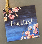 The Kort från Gamla Stans Kreativa kort Grattis by BUTIK KUBIK is a charming 65x65mm card featuring Grattis! in white with pink flowers on a dark blue background, adorned with a rustic jutesnöre loop in the top left corner—ideal as a little presentkort.