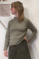 A person with shoulder-length hair and glasses, wearing BUTIK KUBIKs olive-green Kubik sweater waffle and matching skirt of organic fabric, stands sideways against a white wall. A 60s-inspired knit top enhances their outfit while a background wall sign adds visual interest.