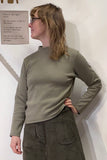 A person with shoulder-length hair and glasses, wearing BUTIK KUBIKs olive-green Kubik sweater waffle and matching skirt of organic fabric, stands sideways against a white wall. A 60s-inspired knit top enhances their outfit while a background wall sign adds visual interest.