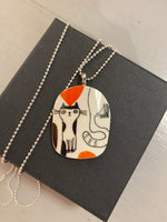 The Saved & Remade Necklace Porslin by BUTIK KUBIK showcases a hand-drawn cat design with orange and brown accents on remade porcelain, hanging from a silver-plated chain. It comes elegantly packaged in a black box.