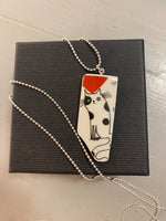 The Saved & Remade Necklace Porslin by BUTIK KUBIK features a black and white cat design on remade porcelain, with a red heart and an elegantly curled tail. It hangs from a silver-plated chain and rests on a closed black box.