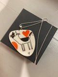 The Saved & Remade Necklace Porslin by BUTIK KUBIK features a heart-shaped pendant with a whimsical cat illustration, accented in black and orange. It comes elegantly attached to a silverpläterad kedja and is presented in a black box.