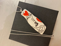 The Saved & Remade Necklace Porslin by BUTIK KUBIK is a unique white pendant made from remade porcelain, featuring a black cat outline and red heart on a silver ball chain, elegantly presented in a closed black box.