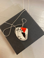 The Saved & Remade Porslin necklace by BUTIK KUBIK showcases a minimalist drawing of a black and white cat on porcelain, accented with a red triangle. It features a silver-plated ball chain and comes in a sleek black box labeled Saved & Remade.