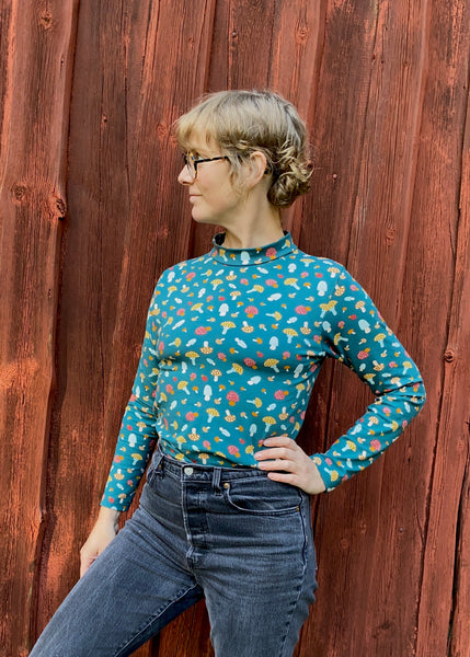 A person confidently gazes to the side, dressed in a blue Kubik sweater mushroom XL by BUTIK KUBIK. The colorful mushroom pattern is made from GOTS certified fabric, paired with dark jeans. A rustic wooden wall with a reddish-brown hue provides warmth to the scene.