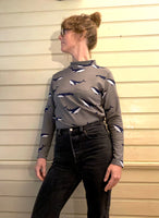 A person with glasses and tied-back hair stands against a wooden wall, wearing the 60s inspired BUTIK KUBIK Kubik sweater whales with long sleeves and whale patterns, paired with dark jeans, looking to the side.