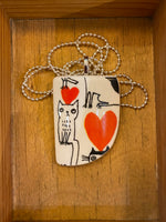 The Saved & Remade Necklace Porslin by BUTIK KUBIK features a wooden frame pendant with black cat illustrations and red heart designs on an återbrukat porslin background, hanging from a silverpläterad kedja.