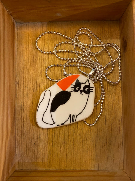 The Saved & Remade Necklace by BUTIK KUBIK features a charming cartoon cat pendant with black spots and an orange party hat, gracefully resting on a wooden surface. The whimsical pendant dangles from a silver-plated chain, adding a playful yet elegant touch to any outfit.