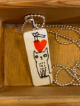 The Saved & Remade necklace Porslin by BUTIK KUBIK is a rectangular pendant in remade porcelain on a silver-plated chain, featuring minimalist cat line art and a red heart. Its primarily white, presented in a wooden box—a perfect piece from the Saved and Remade collection.