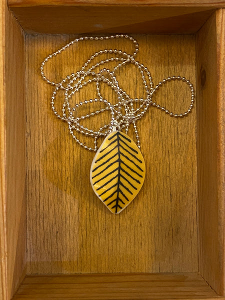 Inside a wooden box lies the Saved & Remade Necklace Porslin by BUTIK KUBIK: a silver-plated chain with small beads and a yellow, leaf-shaped pendant adorned with black lines.