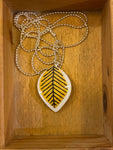 The Saved & Remade Necklace Porslin by BUTIK KUBIK features a leaf-shaped pendant with black and yellow stripes on a silver-plated chain, elegantly presented in a wooden box, celebrating creativity and sustainability.