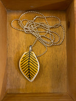 The Saved & remade necklace Porslin by BUTIK KUBIK features a yellow leaf-shaped porcelain pendant with black veins and a silver-plated chain, presented in a wooden box.