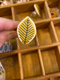 A hand holds a BUTIK KUBIKs Saved & Remade silver-plated knob shaped like a leaf against a yellow background with black veins. Its in a wooden grid organizer with other porcelain knobs, exemplifying Saved & Remade artistry.