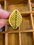 A hand showcasing the Saved & Remade Ring Porslin by BUTIK KUBIK features a teardrop pendant on a yellow background with a black fern pattern, elegantly contrasting against the wooden organizer.