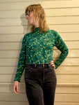 A person with shoulder-length hair and glasses wears the Kubik sweater doodles in green by BUTIK KUBIK, paired with black jeans. They stand against a beige wooden wall, gazing thoughtfully to the side.