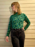 A person with shoulder-length hair and glasses wears the Kubik sweater doodles in green by BUTIK KUBIK, paired with black jeans. They stand against a beige wooden wall, gazing thoughtfully to the side.