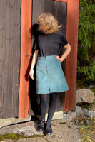 In front of a wooden wall, a person with light brown hair wears a black shirt paired with a stylish Kubik corduroy zip skirt from BUTIK KUBIK, complemented by black tights and shoes. Sunlight casts its glow, while greenery graces the background.