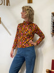 A person with wavy blond hair wears the BUTIK KUBIK Kubik sweater retro 1974, a 60s-inspired floral knit in orange, yellow, and pink, paired with blue corduroy pants. Made in Sweden from organic GOTS cotton.