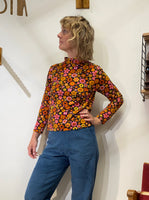 A person with wavy blond hair wears the BUTIK KUBIK Kubik sweater retro 1974, a 60s-inspired floral knit in orange, yellow, and pink, paired with blue corduroy pants. Made in Sweden from organic GOTS cotton.