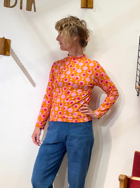 A woman with curly blonde hair stands sideways, wearing the BUTIK KUBIK Kubik sweater popsicle, an orange 60s-inspired knit top with pink and white circles, paired with blue corduroy pants, against a white wall with wooden shelves.
