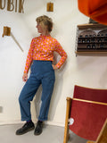 A person with short, curly hair stands indoors wearing the vibrant Kubik sweater popsicle by BUTIK KUBIK, blue pants, and black shoes. They lean against a wall near a jewelry shelf and a red chair.