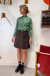 A woman wearing glasses, the BUTIK KUBIK Kubik sweater spring onion, a dark corduroy skirt with front pockets and zipper, black shoes, and rainbow-striped socks stands indoors near a red chair.