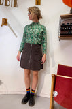 A woman wearing glasses, the BUTIK KUBIK Kubik sweater spring onion, a dark corduroy skirt with front pockets and zipper, black shoes, and rainbow-striped socks stands indoors near a red chair.