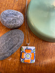 The BUTIK KUBIK Saved & remade necklace Porcelain features an orange and blue flower on upcycled porcelain, hanging from a silver chain, displayed on wood beside two gray stones and a green round object.
