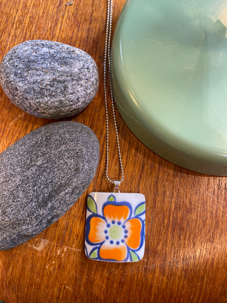 The BUTIK KUBIK Saved & remade necklace Porcelain features an orange and blue flower on upcycled porcelain, hanging from a silver chain, displayed on wood beside two gray stones and a green round object.