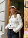 A woman with long blonde hair, wearing the Jumperfabriken cardigan Alida by BUTIK KUBIK, black jeans, and a light-colored cap, stands smiling in a doorway with her hands in her pockets.