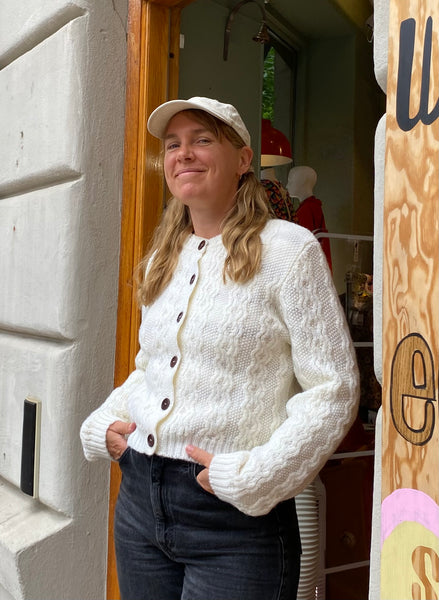 A woman with long blonde hair, wearing the Jumperfabriken cardigan Alida by BUTIK KUBIK, black jeans, and a light-colored cap, stands smiling in a doorway with her hands in her pockets.