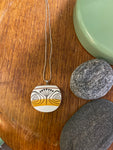 The Saved & Remade Necklace Porcelain by BUTIK KUBIK features a round pendant with a black and yellow abstract tree design on upcycled porcelain, hanging from a silver chain, displayed on wood beside two gray stones and a green accent.