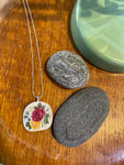 The BUTIK KUBIK Saved & remade necklace Porcelain, featuring a floral design on a silver-plated chain, rests on wood beside two smooth gray stones and a green ceramic dish.
