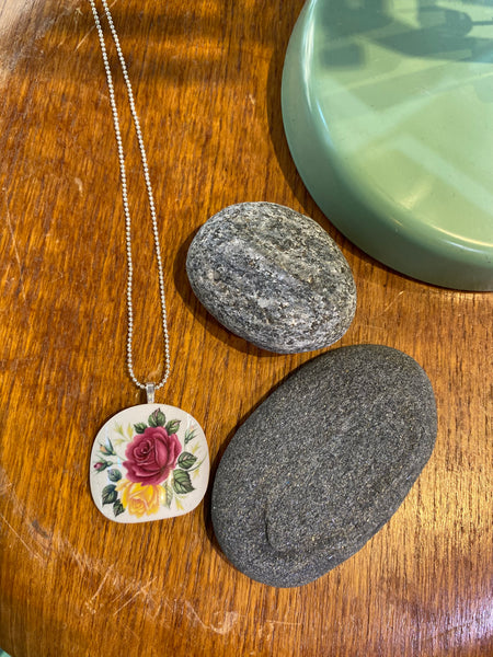The BUTIK KUBIK Saved & remade necklace Porcelain, featuring a floral design on a silver-plated chain, rests on wood beside two smooth gray stones and a green ceramic dish.