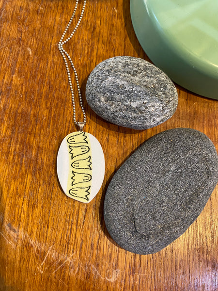 The BUTIK KUBIK Saved & remade necklace Porcelain rests on a wooden surface near two smooth gray stones and a green object in the corner.