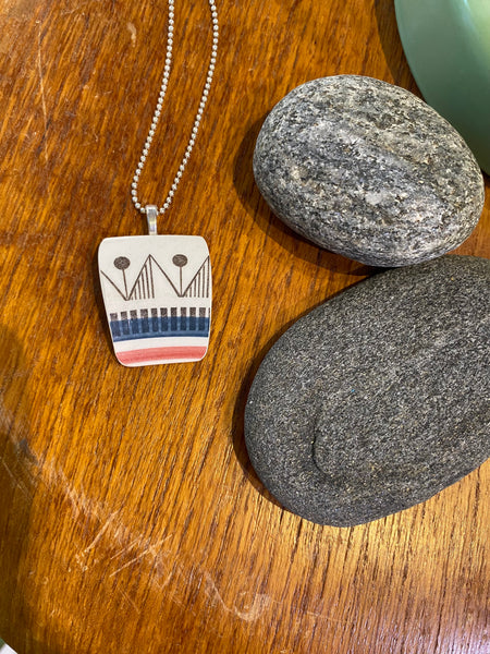 The Saved & remade necklace Porcelain by BUTIK KUBIK features geometric designs from recycled porcelain on a silver-plated chain, displayed on wood beside two smooth gray stones.