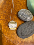 The Saved & Remade Necklace Porcelain by BUTIK KUBIK, featuring a geometric pattern on a silverplated chain, rests on wood beside two smooth gray stones and a green ceramic object.