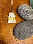 The Saved & Remade Necklace Porcelain by BUTIK KUBIK, featuring a yellow stripe and black line pendant on a silver-plated chain, rests on a wooden surface beside two smooth gray stones.