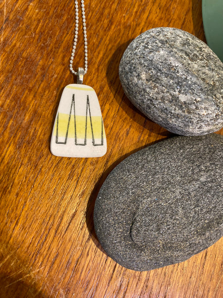 The Saved & Remade Necklace Porcelain by BUTIK KUBIK, featuring a yellow stripe and black line pendant on a silver-plated chain, rests on a wooden surface beside two smooth gray stones.