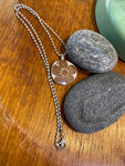 BUTIK KUBIK’s Saved & remade necklace Porcelain, med ett runt hänge av återbrukat porslin med blommönster på silverpläterad kedja, visas på träbredvid grå stenar och ett grönt föremål.