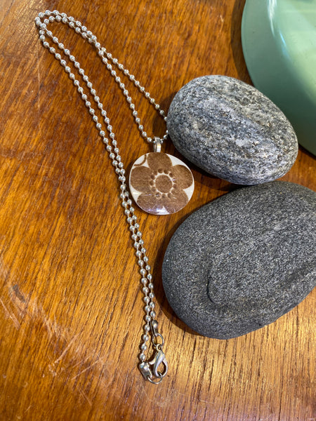 BUTIK KUBIK’s Saved & remade necklace Porcelain, med ett runt hänge av återbrukat porslin med blommönster på silverpläterad kedja, visas på träbredvid grå stenar och ett grönt föremål.