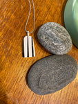 The Saved & remade necklace Porcelain by BUTIK KUBIK, featuring a rectangular pendant with vertical black and white stripes made from återbrukat porslin, rests on wood beside two oval gray stones.