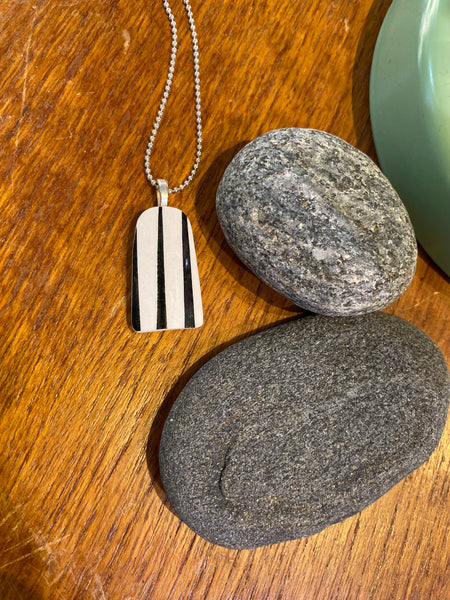 The Saved & remade necklace Porcelain by BUTIK KUBIK, featuring a rectangular pendant with vertical black and white stripes made from återbrukat porslin, rests on wood beside two oval gray stones.