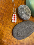 The Saved & Remade Necklace Porcelain by BUTIK KUBIK features a rectangular pendant of recycled porcelain with red and white polka dots, hanging from a silver-plated chain and displayed beside grey stones and green ceramic.