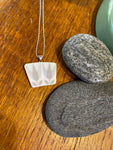 The Saved & Remade Necklace Porcelain by BUTIK KUBIK features a rectangular white pendant of recycled Berså Stig Lindberg porcelain with a leaf motif, hanging on a silver-plated chain alongside gray stones and green sea glass.
